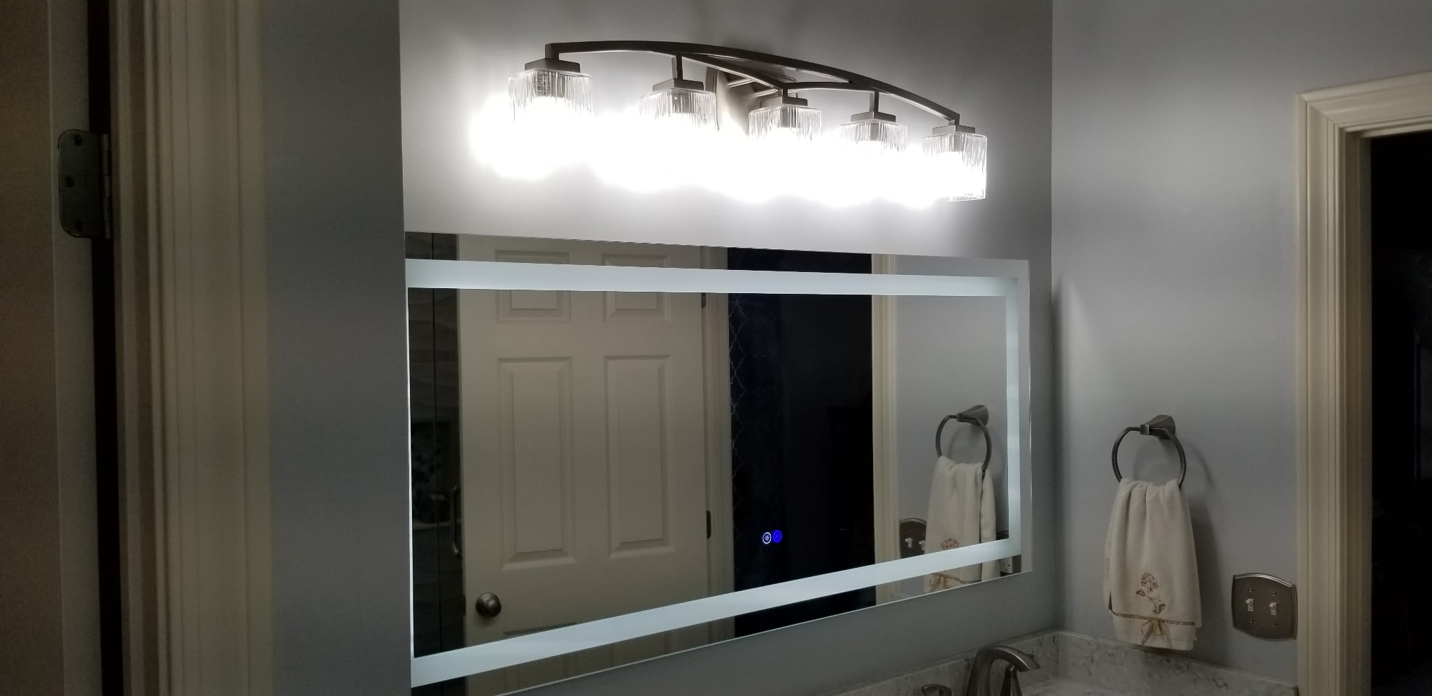 Backlit vanity mirror with arched crystal light bar above marble countertop