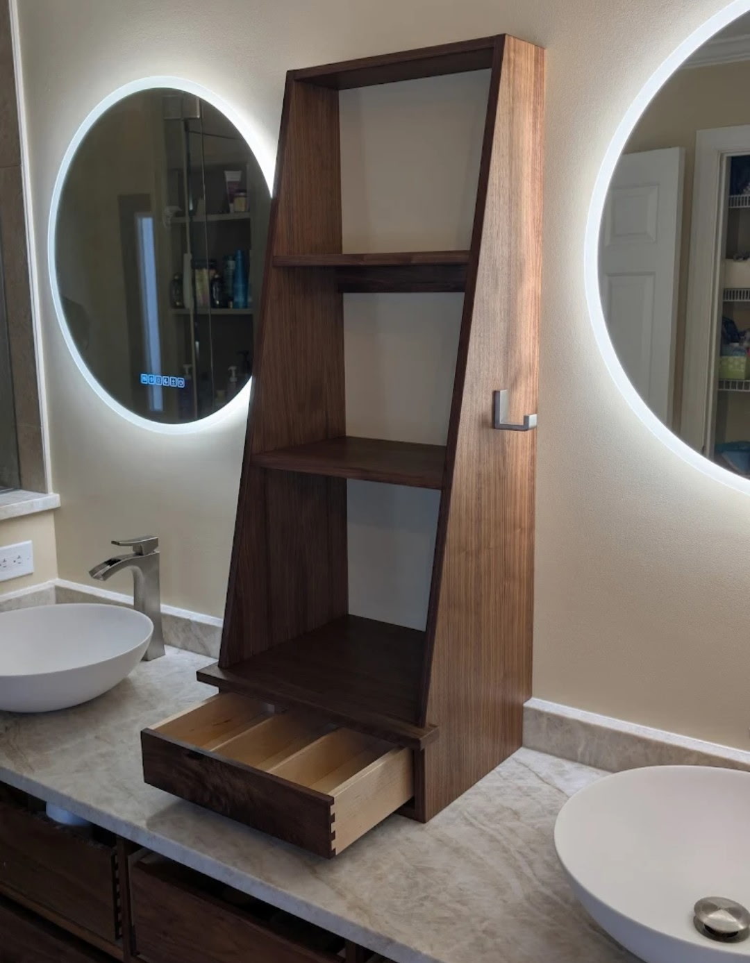 Custom walnut tower shelf unit between backlit round mirrors and vessel sinks