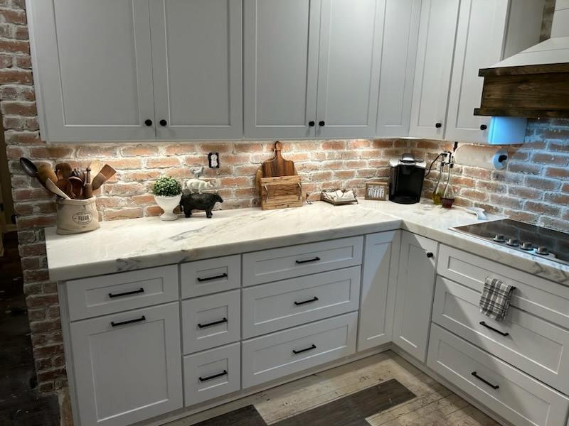 White shaker cabinets with marble countertops and exposed brick backsplash